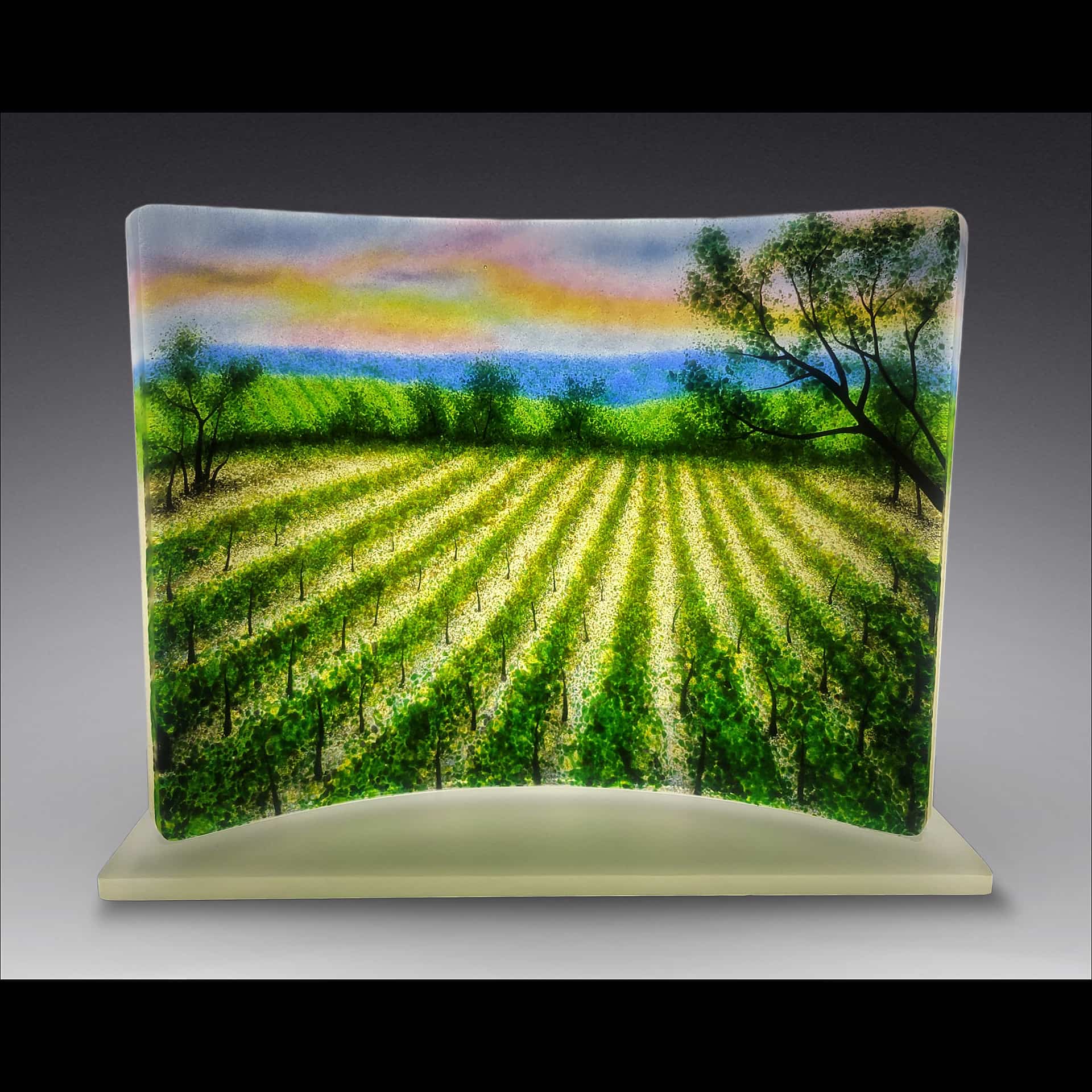 Fused-glass artwork by David Norton showing a sunlit vineyard with rows of bright green grapevines beneath a colorful sky, created with Bullseye clear sheet, colored frit, and Rogue black enamel, fired in a Jen-Ken Profusion 26 kiln.