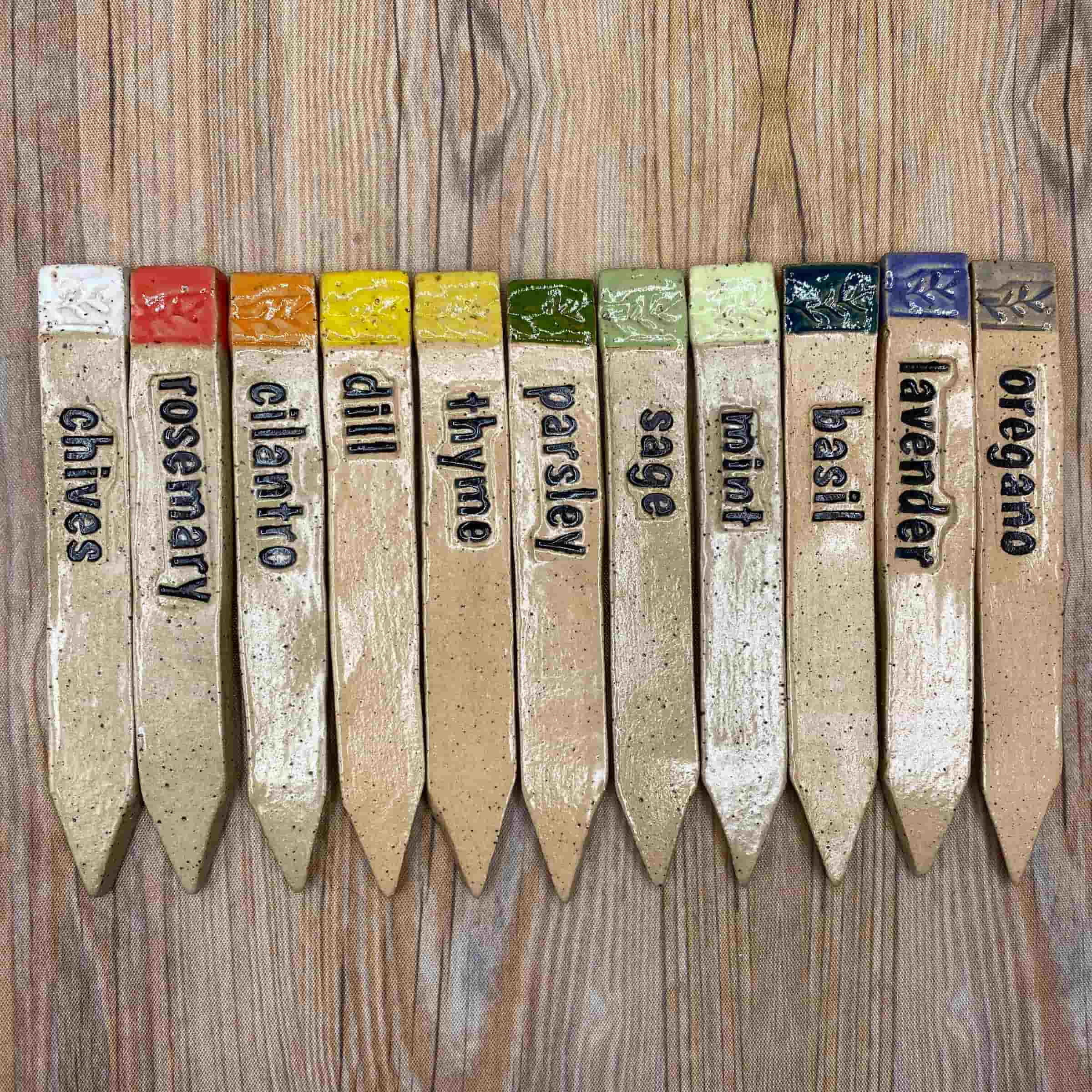 A set of handmade ceramic herb markers arranged in a row on a wood-textured surface. Each marker is labeled with a herb name—chives, rosemary, cilantro, dill, thyme, parsley, sage, mint, basil, lavender, and oregano—and topped with colorful glazed squares in reds, yellows, greens, and blues.