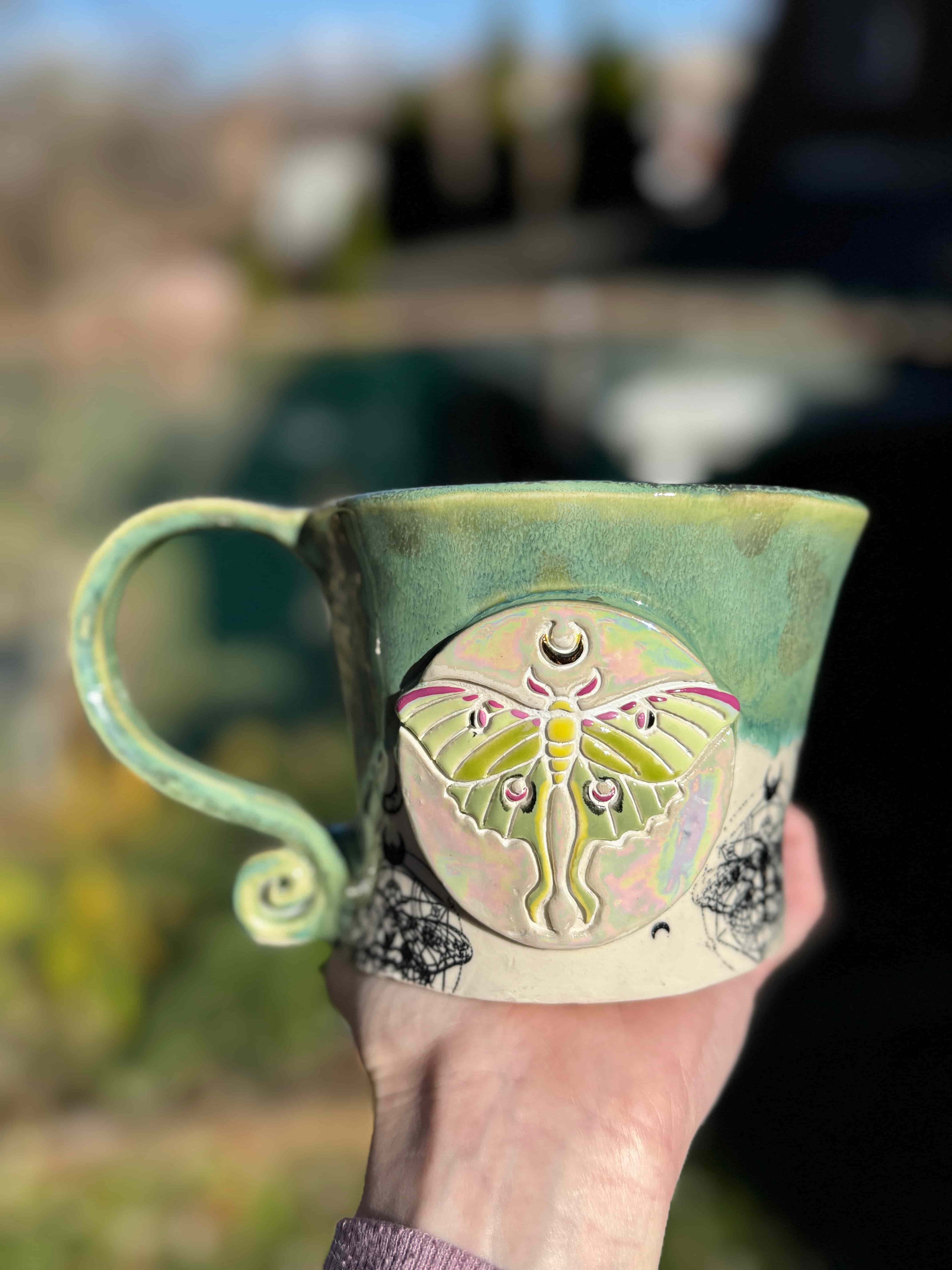 A handcrafted ceramic mug with a soft green glaze and a sculpted luna moth medallion on the front. The moth features iridescent pastel tones and fine detailing, while the mug’s handle curls into a whimsical spiral. Held outdoors with a softly blurred background.