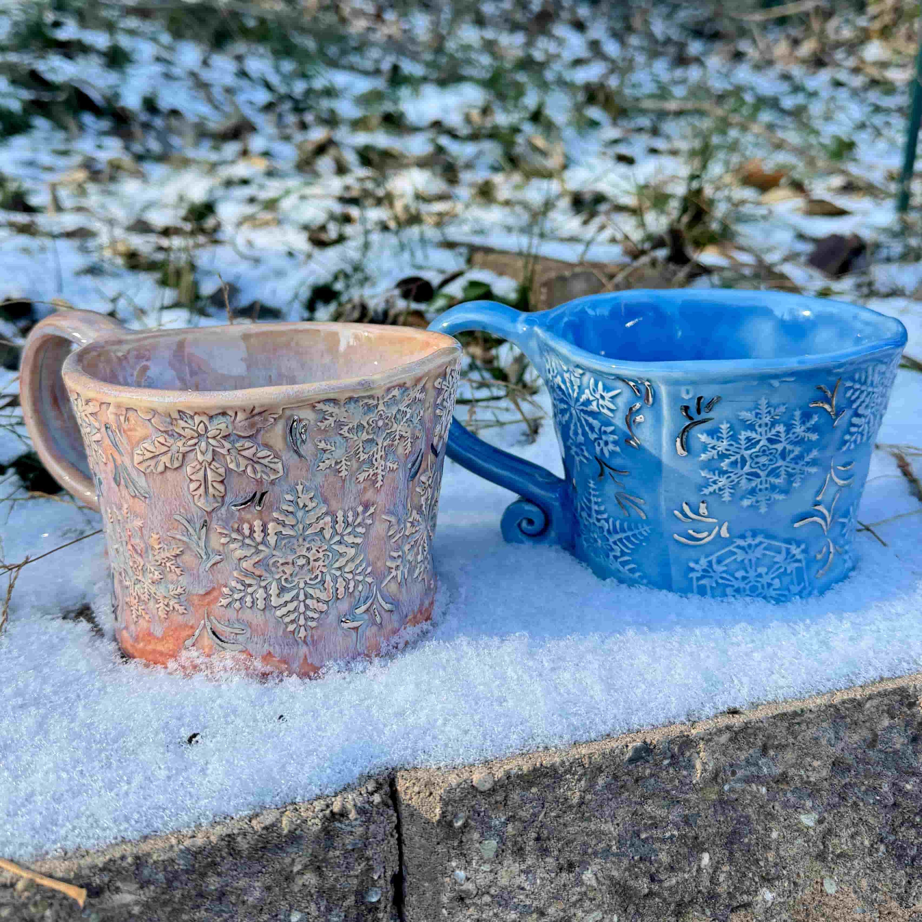 Two textured ceramic mugs with intricate raised snowflake patterns, displayed outdoors in the snow. One mug is a soft blush-pink glaze with white accents, and the other is a vibrant icy blue with shimmering details. Both feature expressive, sculpted handles.