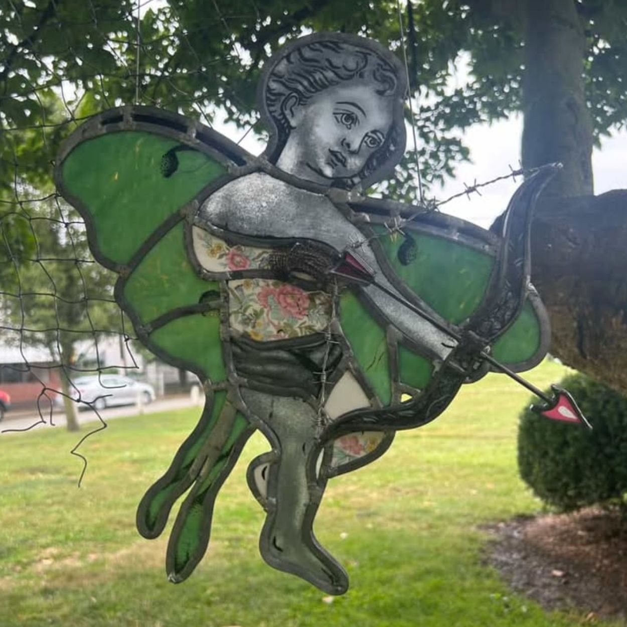 Stained glass artwork by Cosmik Jest depicting a winged, human-like figure with green wings and a grayscale face, holding a bow and arrow. The body incorporates patterned glass and layered textures, with visible lead lines outlining the figure. The piece is photographed outdoors with trees and grass in the background.