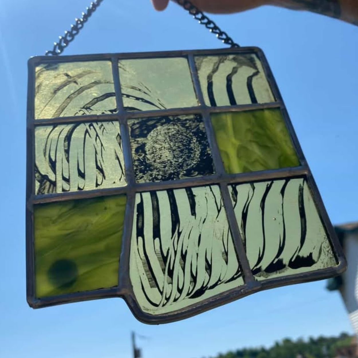 Abstract stained glass panel by Cosmik Jest featuring a grid of rectangular sections filled with pale green and olive glass. Several panels include black, organic linework resembling melting or flowing shapes. The piece hangs from a chain against a blue sky, emphasizing transparency and texture.