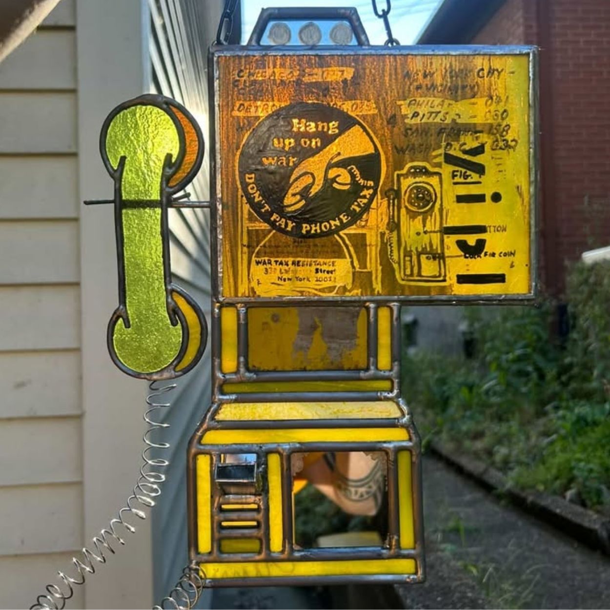 Stained glass sculpture by Cosmik Jest depicting a vintage wall-mounted telephone in yellow and amber glass tones, with a green handset connected by a coiled wire. The glass includes layered imagery, text, and graphic elements, framed with dark lead lines. The piece hangs outdoors against a house and garden background.