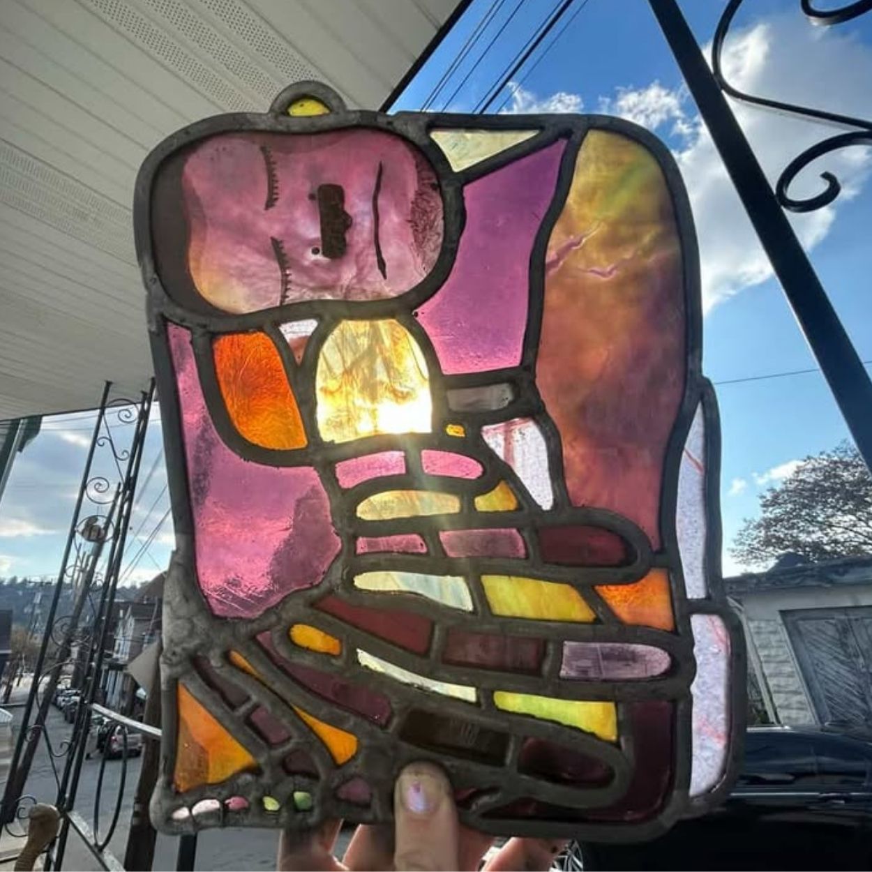 Abstract stained glass figure by Cosmik Jest composed of pink, purple, orange, and yellow glass segments outlined in dark lead. The figure appears reclined or resting, with simplified facial features and layered color transitions. The piece is held up outdoors, with sunlight shining through the glass and a residential street visible behind it.