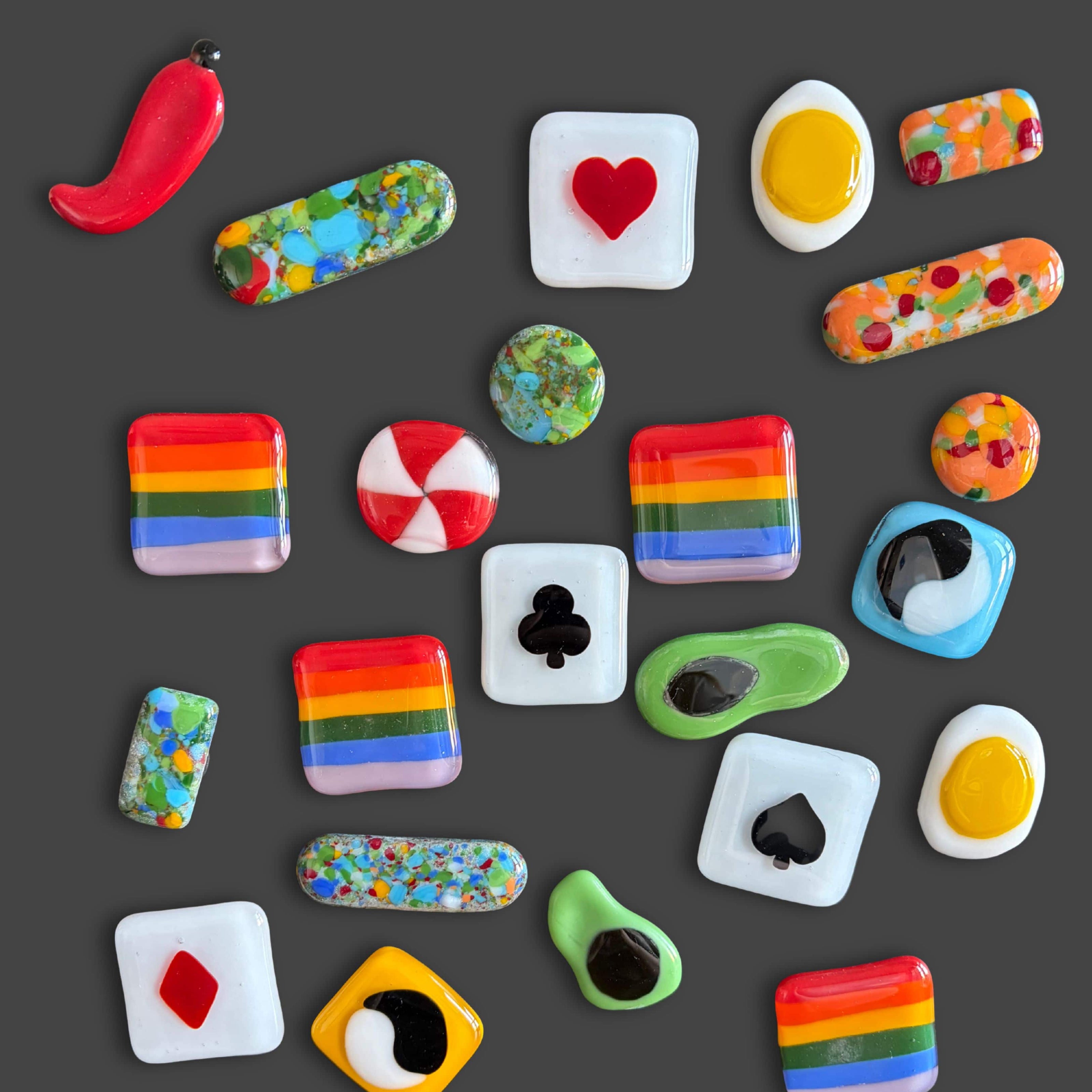 Colorful fused glass trinkets by Grace Pratchett of Patch of Glass, including rainbow-striped squares, heart and club tiles, pill-shaped pieces with multicolored frit, and playful abstract glass designs on a dark background.