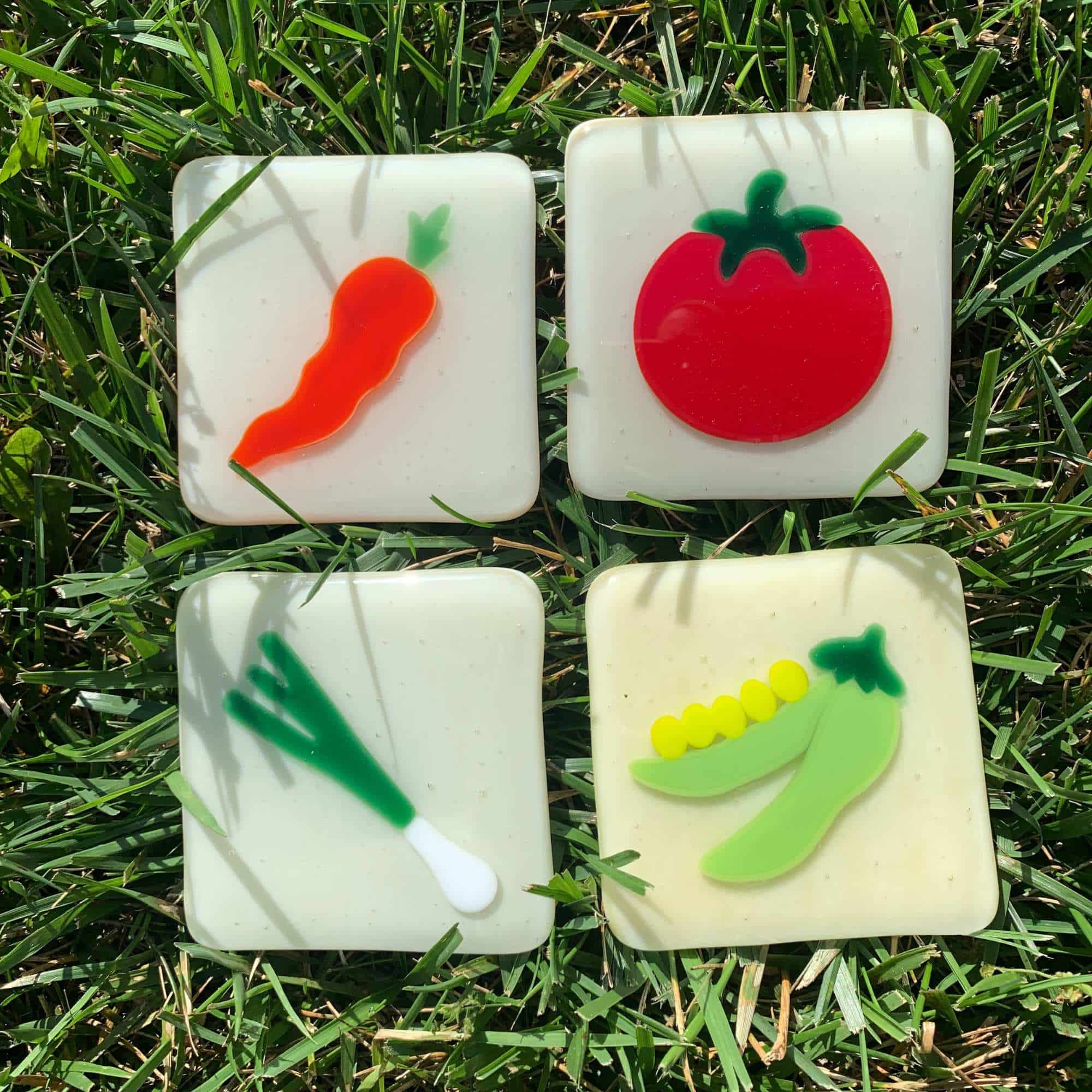 Fused glass vegetable tiles by Grace Pratchett of Patch of Glass, featuring a carrot, tomato, green onion, and pea pod designs on soft white square bases, photographed outdoors on grass.