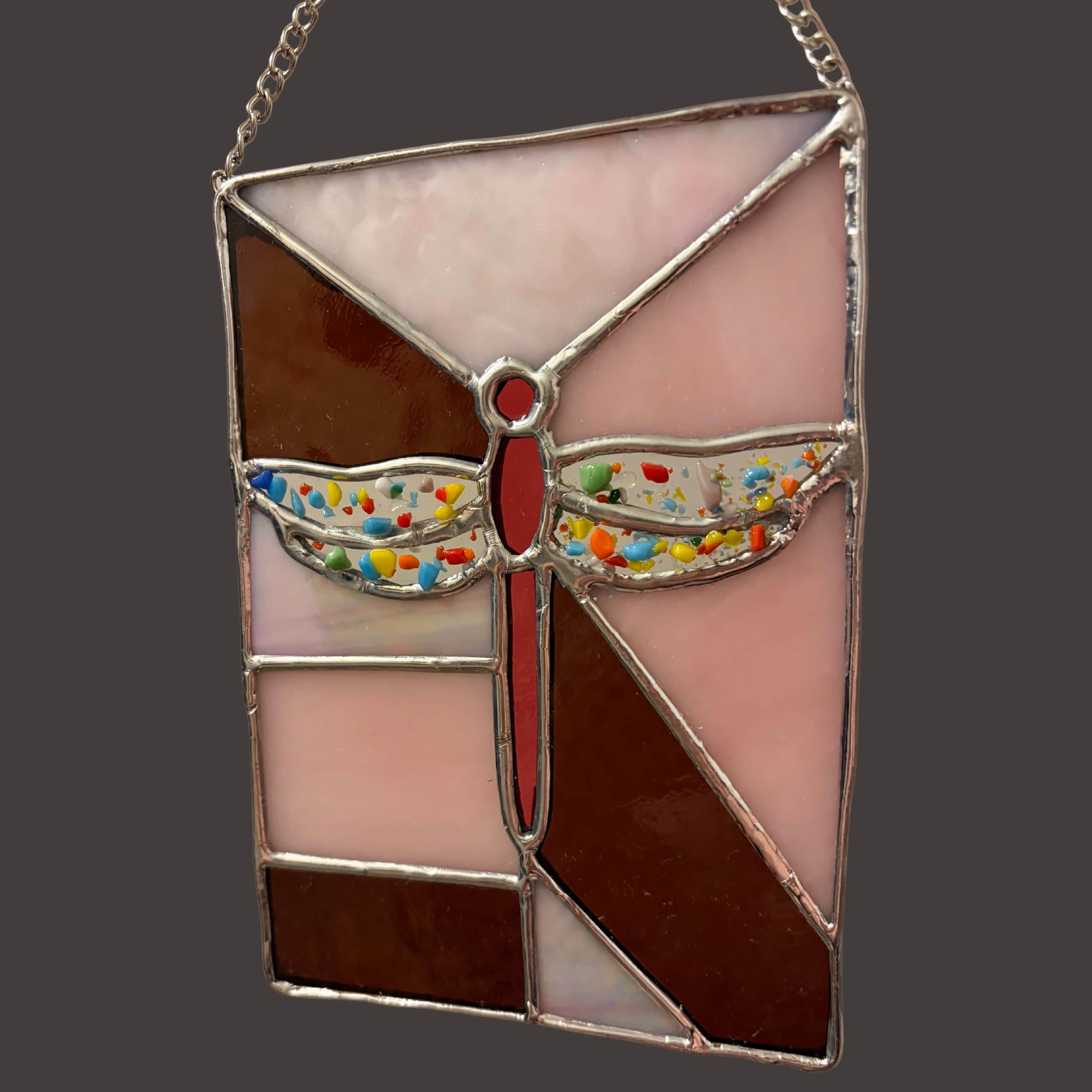 Stained glass dragonfly panel by Grace Pratchett of Patch of Glass, designed with geometric pink and brown glass sections, a red dragonfly body, and multicolored frit wings, hanging against a dark background.
