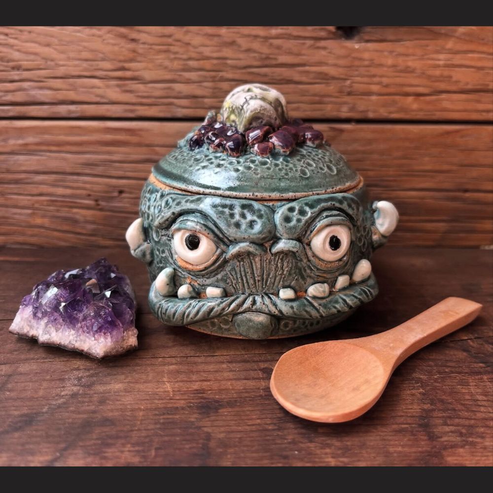 Handmade ceramic creature bowl by Pickled Aliens Pottery featuring a textured green glaze, wide eyes, and sculpted facial details, shown with a wooden spoon and crystal accent; fired in a Jen-Ken electric kiln.