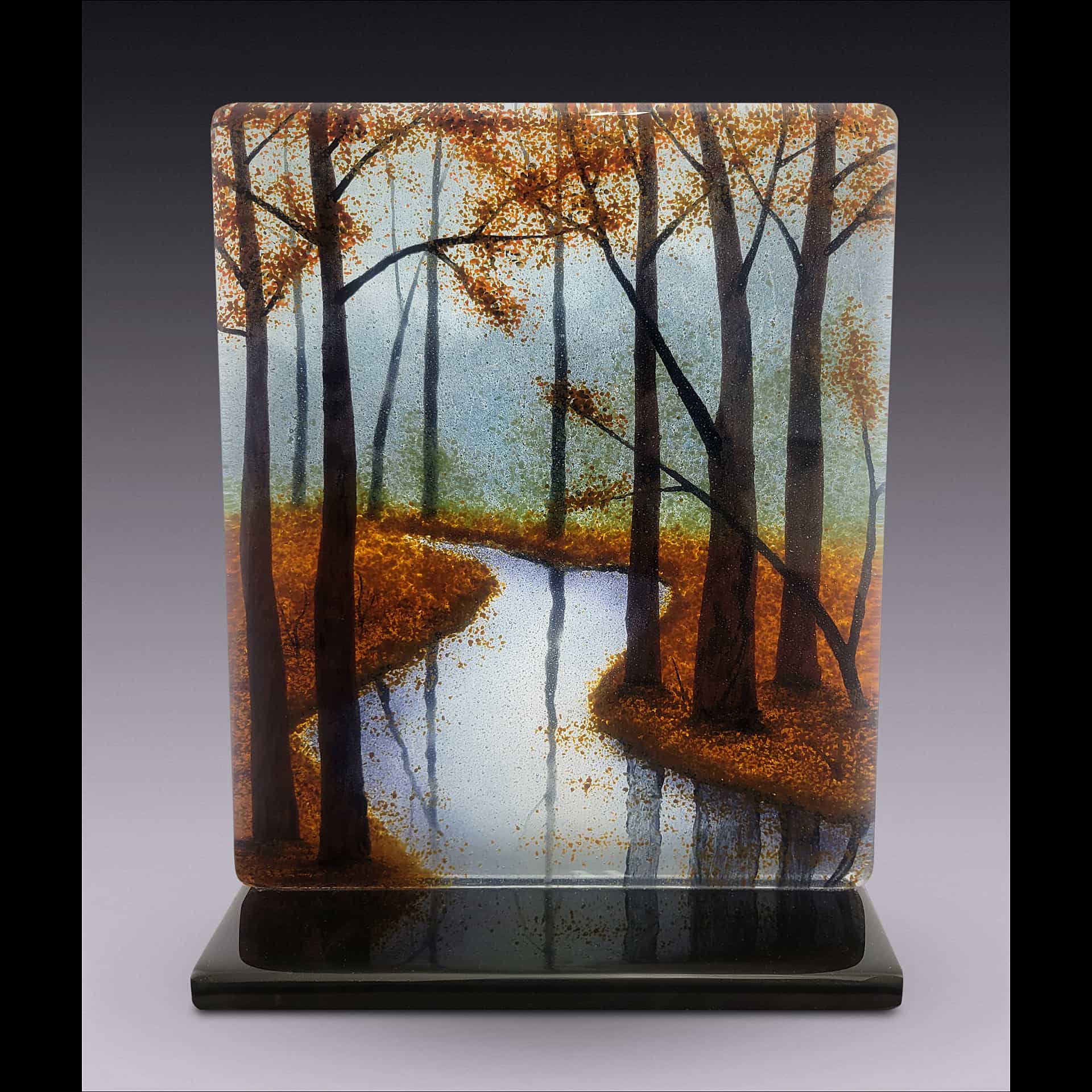 Fused-glass landscape by David Norton showing an autumn forest with vibrant orange foliage surrounding a reflective winding stream, made with Bullseye clear sheet, colored frit, and Rogue black enamel, fired in a Jen-Ken Profusion 26 kiln.