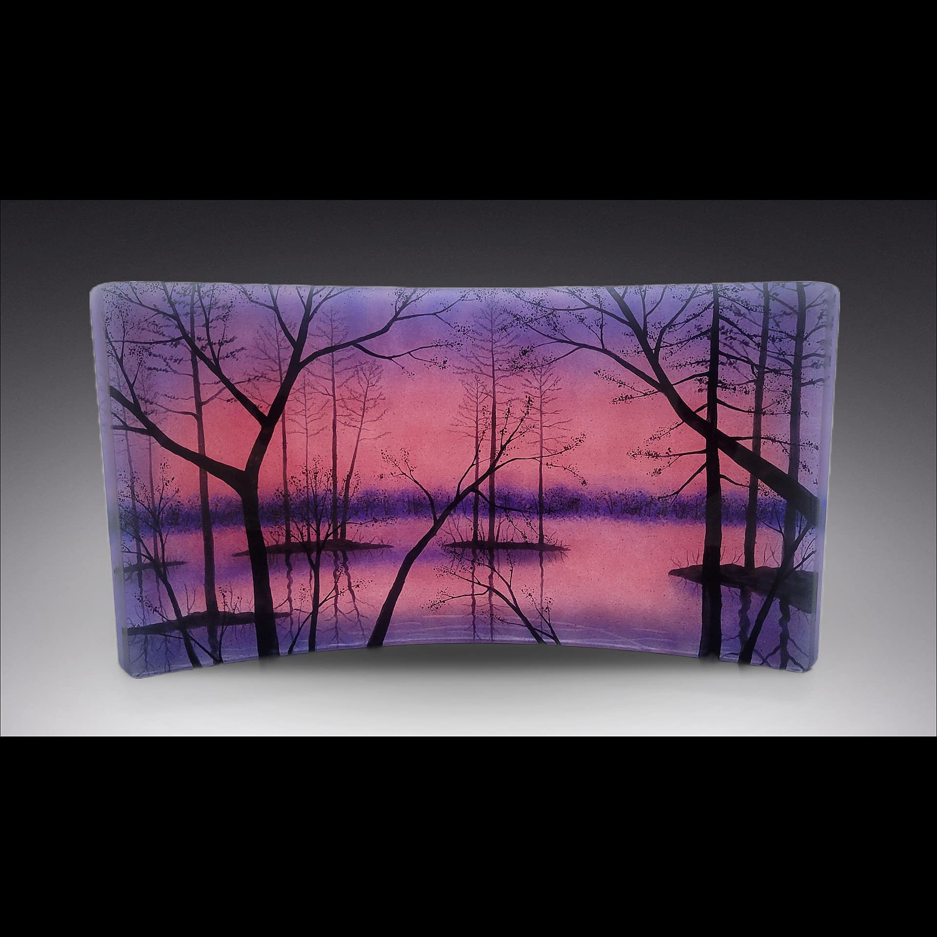 Curved fused-glass panel by David Norton featuring silhouetted trees against a vivid pink-purple sunset over a calm lake, crafted using Bullseye clear sheet, colored frit, and Rogue black enamel, fired in a Jen-Ken Profusion 26 kiln.